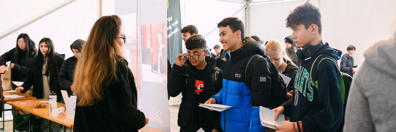 Schüler*innen stehen an Tischen und sprechen mit einer Beraterin in einem Zeltinneren.