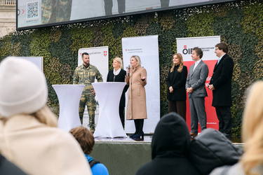 Politische Veranstaltung mit mehreren Personen am Rednerpult, darunter ein Soldat und Vertreter der Regierung.